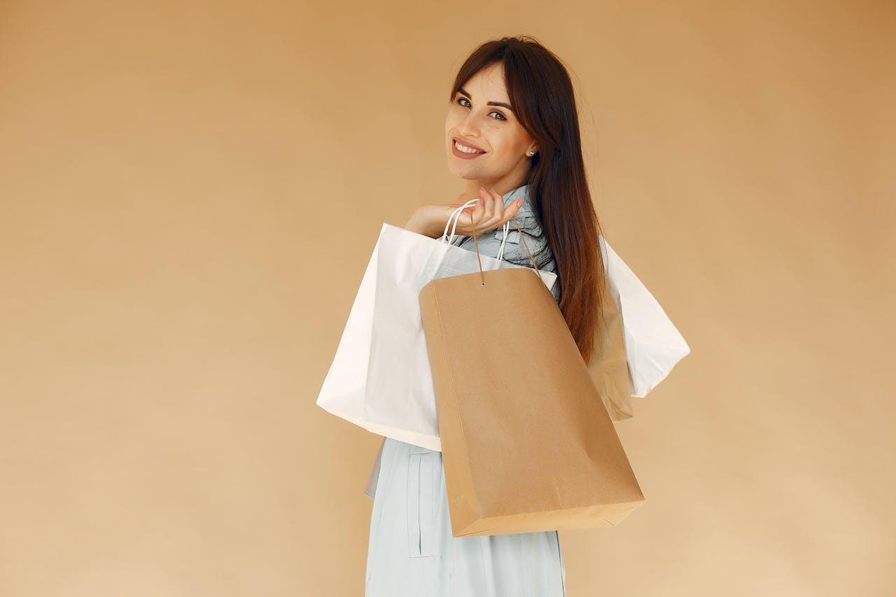 Side view of happy female shopper carrying paper bags while looking at camera over shoulder and standing isolated on brown background